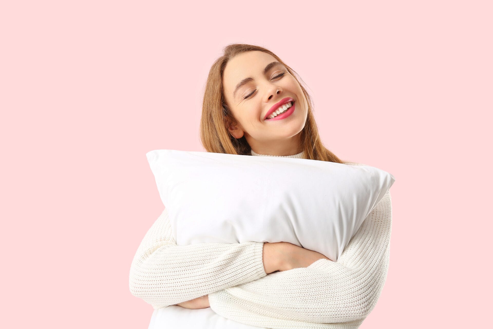 A smiling woman in a white sweater hugs a non-toxic pillow against a pink background, showcasing the comfort and safety of 2025 pillows.
