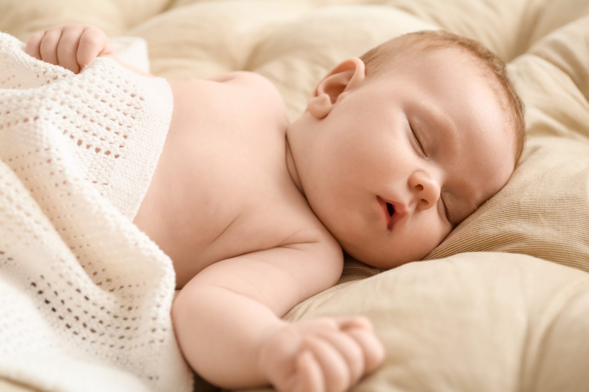 A sleeping baby lies on a soft beige blanket, cuddled among baby pillows, partially covered with a white crocheted blanket.