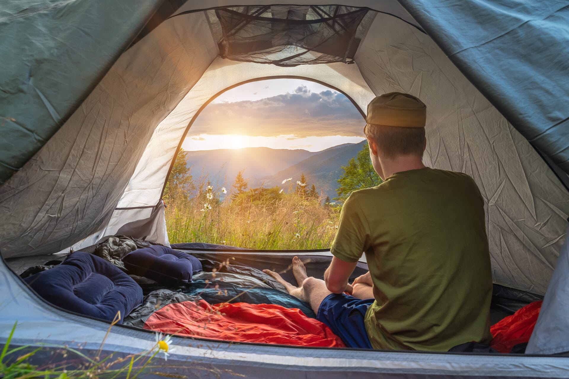 A person sits comfortably in a tent, best camping pillow in hand, gazing out at a breathtaking mountain landscape bathed in the warm hues of sunset. Surrounding them is an array of essential camping gear, promising a cozy night ahead amidst nature's grandeur.