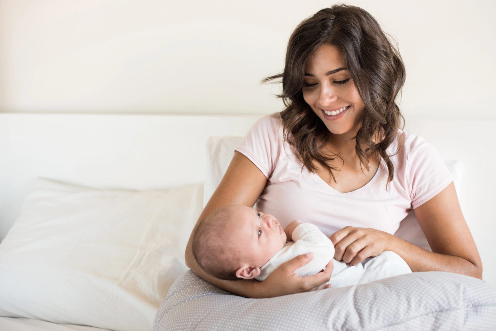 A woman in a pink shirt smiles while holding a baby wrapped in a blanket, comfortably sitting on a bed with white pillows and bedding, showcasing the best nursing pillow for ultimate support.